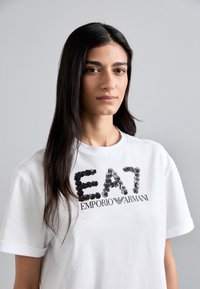 Bijela pamučna majica s kratkim rukavima koja ima iskrice na "EA7" logo-u i crni tekst "EMPORIO ARMANI" ispod. Okrugli izrez.