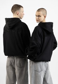 Svart oversized hettegenser med slitte detaljer bak, kombinert med lysegrå baggy jeans med baklommer.
