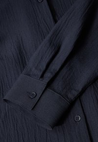 Navyblauwe shirtmanchet van een gestructureerde stof, met een ronde zoom en een enkele zwarte knop. Zichtbare kreuken voegen diepte aan het ontwerp toe.