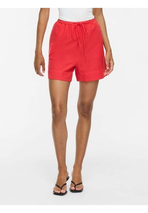 Personne portant un short à cordon rouge vif et des sandales noires à bouts ouverts, debout devant un fond blanc uni.