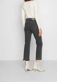 Mujer vistiendo un suéter de canalé color crema, jeans negros cortos y deshilachados, y botines de tacón color crema, de pie contra un fondo claro y liso.