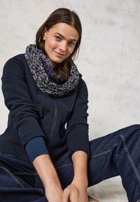 Sort-hvide mønstrede infinity-sjal båret over en marinblå zip-up jakke, parret med mørke denimbukser. Blødt stof i tekstur, cirkulært design.