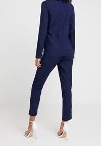 Žena stojící s otočeným zády, oblečená ve štíhlém námořnickém obleku a bílých vysokých sandálech na pozadí čistě bílé barvy.