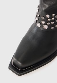 Botas de cuero negro con punta cuadrada y tachuelas decorativas plateadas alrededor del tobillo sobre un fondo blanco.