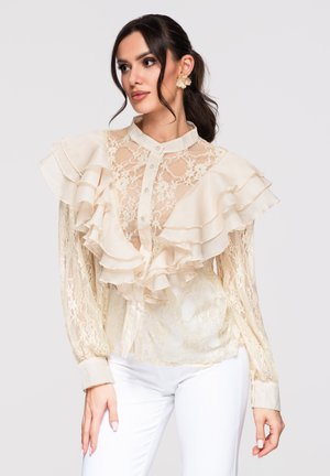 Nainen yllään läpikuultava kermaisen värinen pitsipaita, jossa on kerrokselliset röyhelöt harteilla, ja valkoiset housut, katsoo sivulle hiukset sidottuna taakse.