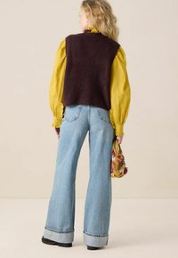 Gebreide bordeaux vest over een gele T-shirt met lange mouwen, gecombineerd met lichtblauwe flared jeans en een kleurrijke handtas.