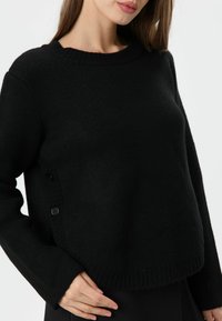 Schwarzer Strickpullover mit rundem Halsausschnitt, strukturierter Stoff und gerippten Bündchen. Mit einer Knopfdetail an der Seite. Bequeme und entspannte Passform.