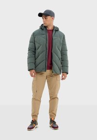 Veste verte matelassée avec un design matelassé diagonal, casquette grise, chemise bordeaux, pantalon cargo beige et baskets multicolores.
