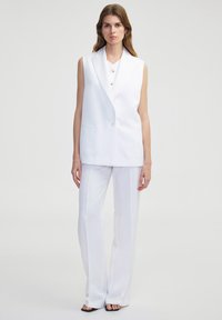Witte mouwloze blazer met een V-hals en sluiting met één knop, gecombineerd met witte op maat gemaakt broeken, wat een monochromatische outfit creëert.