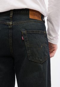 Οπίσθια όψη ατόμου π ου φοράει σκούρο μπλε τζιν Levi's με ορατό δερμάτινο λογότυπο και κόκκινη ταμπέλα, σε συνδυασμό με ένα λευκό μακρυμάνικο πουκάμισο.