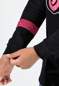 Felpa nera con un accento di paillettes rosa sulla manica. Presenta polsini a costine e una vestibilità comoda. Ingrandimento di una mano che regola la manica.