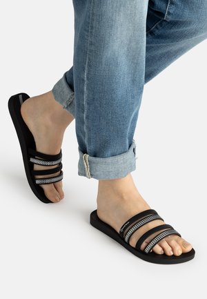 Person trägt hochgekrempelte blaue Jeans und schwarze Slip-on Sandalen mit drei Riemen und silbernen Kettendetails vor weißem Hintergrund.