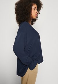 Maglietta a maniche lunghe color blu navy con taglio morbido, caratterizzata da un scollo a V e una texture morbida e liscia. Abbinata a pantaloni di colore chiaro.