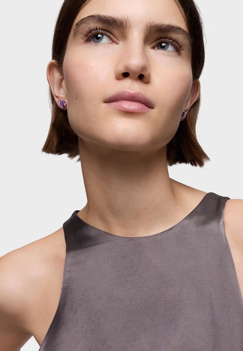 Orecchini a forma di punto viola adornano le orecchie di un modello. Indossa un top senza maniche di satin grigio scuro con una texture liscia e scollatura rotonda.