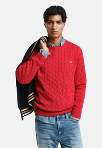 Rode kabelgebreide trui met een ronde halslijn, gecombineerd met een lichtblauw denimhemd en blauwe versleten jeans. Zwart-wit gestreepte jas over de schouders gehangen.