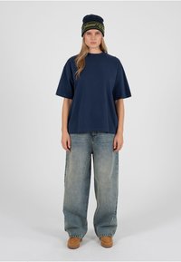 Camiseta de algodón azul marino con mangas cortas, combinada con vaqueros de mezclilla descoloridos y sueltos y botas marrones hasta el tobillo; gorro de punto a la moda con detalles verdes.