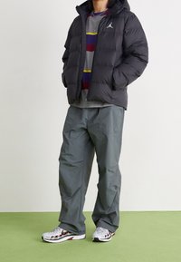 En pufferjacka i svart, grå tröja med färgade ränder, grå byxor och vita sneakers med röda accenter, stående på grönt golv.