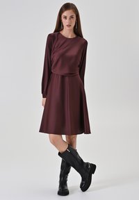 Bordeauxrotes Kleid mit langen Ärmeln und drapiertem Oberteil, knielanger Rock, kombiniert mit knielangen schwarzen Stiefeln mit Reißverschluss und profilierter Sohle.