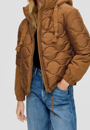 Kvinde iført en lysebrun quiltet jakke med hætte og lynlås, kombineret med blå jeans, stående med den ene hånd i lommen.