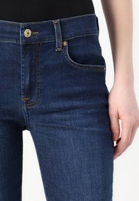 Jeans in denim blu scuro con un design aderente, dotati di tasca frontale e cuciture a contrasto. Dettagli in metallo con bottone in vita.