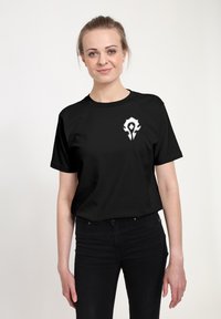Žena se světlými vlasy staženými do culíku nosí černé tričko s bílým stylizovaným emblémem na levé hrudi, v kombinaci s černými kalhotami na jednobarevném pozadí.