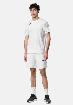 Jeune homme debout portant une tenue de sport blanche avec des logos Kappa noirs, des baskets noires et des chaussettes blanches contre un fond clair uni.
