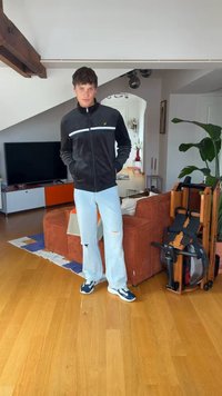 Junger Mann in schwarzer Jacke und zerrissenen hellen Jeans steht in einem modernen Wohnzimmer mit braunem Sofa und einem Rudergerät neben einer Pflanze.