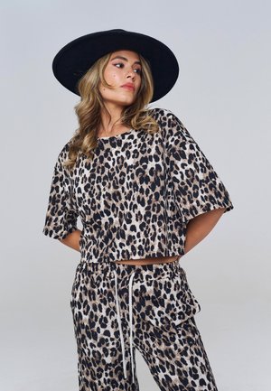 Femme portant un chapeau noir à large bord et un haut et un pantalon amples assortis imprimés léopard, posant les mains dans le dos sur fond gris.