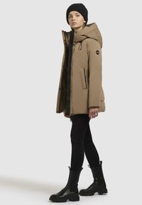 Giacca parka khaki con grande cappuccio, zip frontale e tasche laterali, abbinata a leggings neri e scarpe alla caviglia. Tessuto liscio, stile casual.