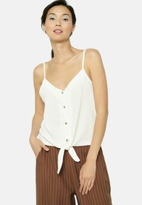 Biely, bezrukávový top s výstrihom do V, predným zapínaním na gombíky a detailom v podobe uzla na hemline. Tkanina má textúrovaný, rebrovaný dizajn.
