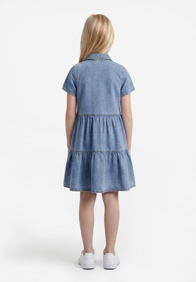 Ξανθό κορίτσι που στέκεται με την πλάτη φορώντας ένα μπλε τζιν φόρεμα με κοντά μανίκια και λευκά αθλητικά παπούτσια σε λευκό φόντο.