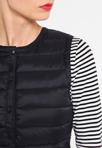 Närbild av en person som bär en svart vadderad väst över en svartvit randig långärmad skjorta, med starkt röd läppstift synligt.