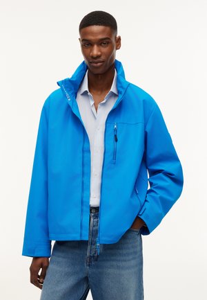 Mand iført en knaldblå lynlåstrøje over en lyseblå skjorte med krave og blå jeans, stående mod en ensfarvet hvid baggrund.