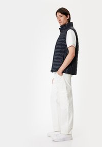 Navy quilted vest with high collar, front zipper, and side pockets, paired with white cargo pants and white sneakers. Smooth, lightweight material.