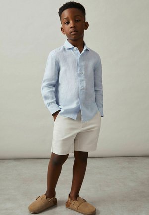 Nuori poika seisoo käsineen taskuissa, päällään vaaleansininen nappipaita, beiget shortsit ja vaaleanruskeat slip-on-kengät.