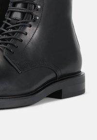 Botín de cuero negro hasta el tobillo con acabado mate, puntera redonda y detalle de cordones. Suela robusta con un ligero tacón y sutiles acentos de costura.