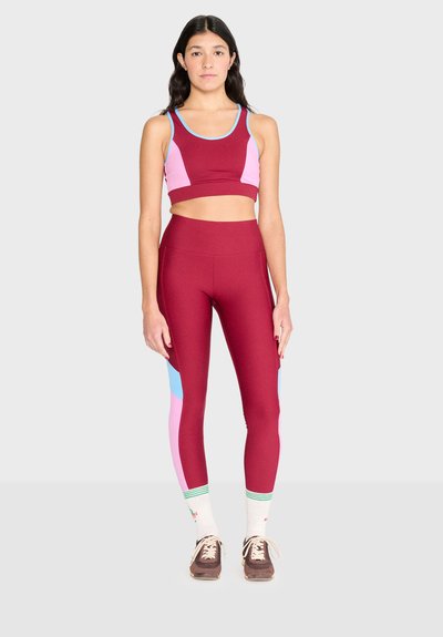 Femme debout portant un brassard de sport à blocs de couleurs bordeaux, rose et bleu, des leggings, des chaussettes blanches et des baskets marron.