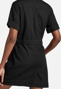 Kvinna i en kortärmad svart klänning med uppvikta ärmar och elastisk midja, sedd bakifrån mot en helt vit bakgrund.