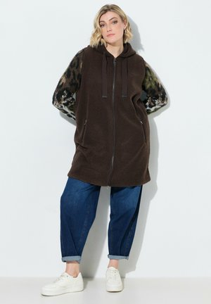 Kvinde poserer mod en hvid væg iført lang brun hættevest med lynlås over leopardmønstrede ærmer, blå manchetjeans og hvide sneakers.