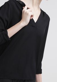 Zwarte V-hals blouse met driekwartmouwen, gladde textuur en een ontspannen pasvorm. Verfraaid met subtiele stikdetails.