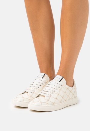 Baskets basses blanches avec des motifs géométriques beiges et des détails de logo, portées pieds nus sur un fond uni.