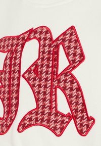 Felpa rossa e bianca con una grande lettera "R" tessuta e ricamata in un motivo houndstooth, bordata da cuciture rosse su uno sfondo bianco.