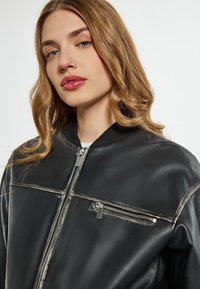 Giacca bomber in pelle nera con cerniera anteriore, collo a coste e tasca sul petto con zip e accessori in metallo argentato.