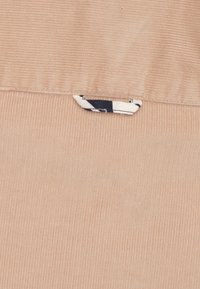 Zachtroze corduroy stof met verticale ribbels. Een klein wit label met een marineblauw patroon is aan de linkerkant weergegeven.