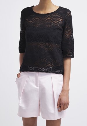 Femme portant un haut en dentelle noire à manches 3/4 et un short plissé taille haute rose clair, debout avec une main dans la poche.