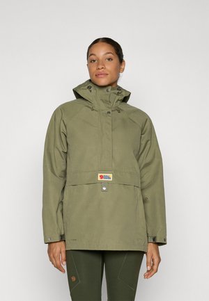 Woman wearing olive green hooded jacket and matching pants, standing against plain light gray background, facing forward.