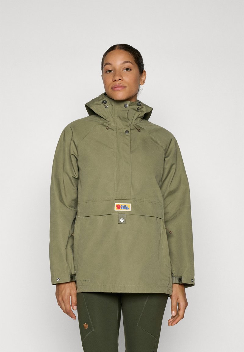 Vrouw in een olijfgroene hooded jacket en bijpassende broek, staand tegen een effen lichtgrijze achtergrond, recht vooruit kijkend.