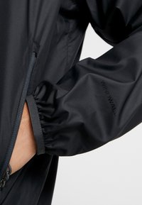 Musta tuulenpitävä takki, jossa on joustavat hihansuut, vetoketjukiinnitys ja tekstuuri materiaali. Brändin "WINDWALL" logo näkyy hihassa.