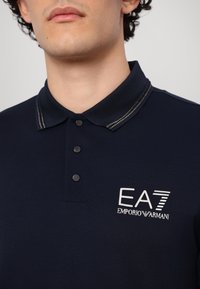 Polo shirt bleu foncé avec un tissu texturé, patte de boutonnage à trois boutons et col rehaussé d'or. Présente un logo blanc sur la poitrine gauche.