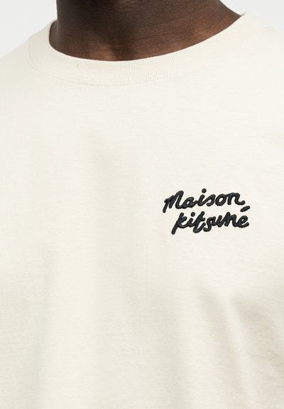 Κοντινό πλάνο ενός μπεζ πουλόβερ με στρογγυλή λαιμόκοψη και μαύρη κεντημένη επιγραφή "Maison Kitsuné" στο αριστερό στήθος.